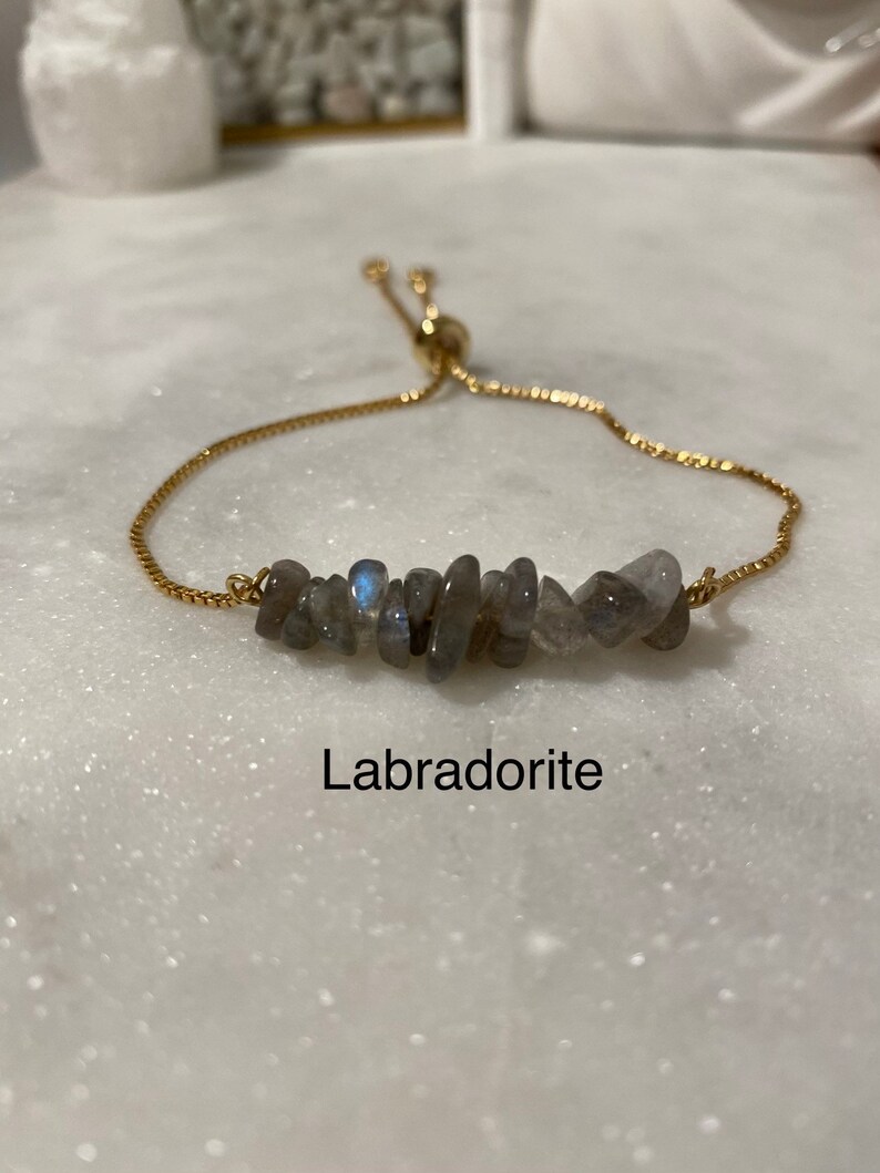 May include: A gold chain bracelet with a slider clasp and a row of grey labradorite stones. The bracelet is on a white surface and the word "Labradorite" is written below the bracelet.