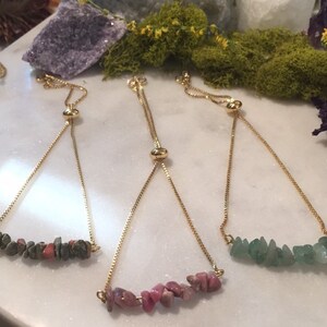 May include: Three adjustable bracelets with gold chains and different colored gemstone beads. The bracelets feature a slider bead for adjusting the length. The beads are green, pink, and a mix of brown and pink.