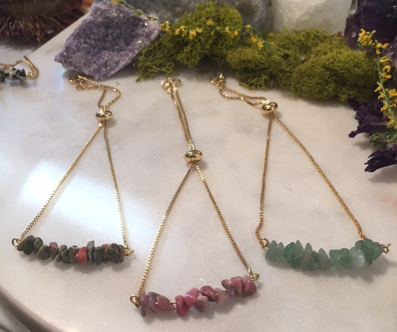 May include: Three adjustable bracelets with gold chains and different colored gemstone beads. The bracelets feature a slider bead for adjusting the length. The beads are green, pink, and a mix of brown and pink.