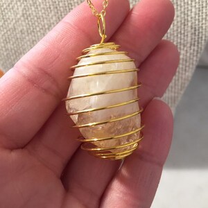 Könnte beinhalten: Ein Anhänger aus goldfarbenem Drahtkäfig mit einem klaren Quarzkristall im Inneren. Der Anhänger hängt an einer goldenen Kette.