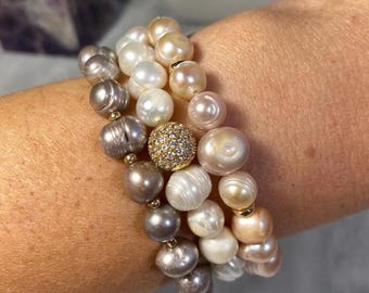Puede incluir: Tres pulseras de perlas en tonos blanco, crema y gris. Una pulsera presenta una cuenta dorada incrustada con pequeñas piedras brillantes. Las perlas son de diferentes tamaños y formas, creando un aspecto único y elegante.