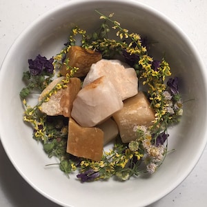 Puede incluir: Un bol blanco lleno de pequeños cristales toscamente cortados, de color marrón claro y beige, rodeados de flores secas amarillas y moradas.