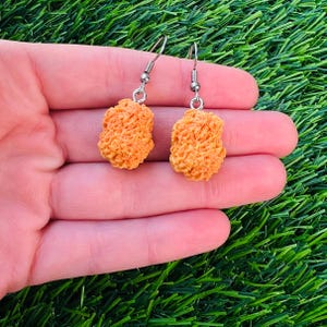 May include: A pair of novelty earrings featuring a fried chicken nugget design. The earrings are a light orange color with a textured surface, suspended from silver-colored fish hook ear wires. The earrings are displayed against a green background.
