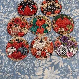 May include: Eight circular coasters featuring diamond art designs of pumpkins in various colors including red, orange, white, green, and purple. Each coaster displays a unique arrangement of pumpkins, some with checkered patterns, bows, or floral accents.