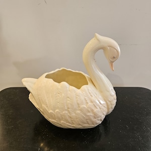 May include: A cream-coloured ceramic swan planter featuring a glossy finish and detailed moulded feather textures on the body. The swan has a long, curved neck with a downward-facing head, a small orange beak, and a hollow opening on its back.