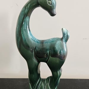 May include: A standing deer figurine crafted from glossy ceramic with a deep emerald green glaze. The sculpture features a long, curved neck, pointed ears, and a short tail, with darker shaded areas highlighting the form and a textured base.