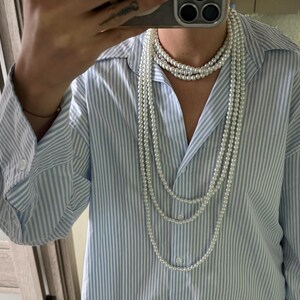 May include: Multiple strands of white pearl necklaces cascading down over a light blue and white striped button-down shirt. The pearls vary in length, creating a layered effect. The shirt has long sleeves.