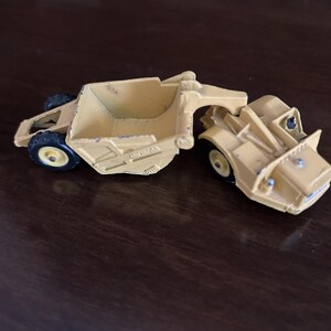 May include: A yellow toy construction grader with black wheels. The grader is made of metal and has a detailed design, including a blade and a cab. The toy is likely a vintage collectible.