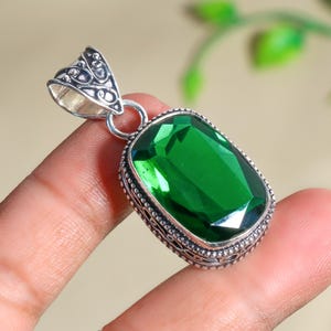 Puede incluir: Un colgante de plata con un diseño adornado presenta una gran piedra preciosa verde esmeralda facetada.