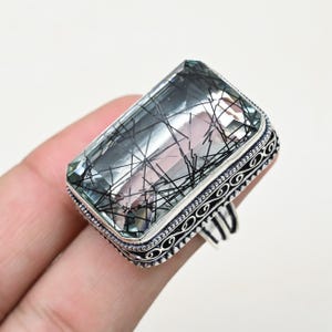 May include: A rectangular gemstone ring with a clear, light green stone featuring black inclusions. The ring has a silver band with an ornate, patterned design around the stone. The ring is held by a person's fingers.