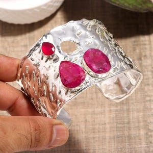 Puede incluir: Un brazalete ancho de plata con una superficie martillada y texturizada, adornado con tres piedras preciosas irregulares de color rojo rosado pulido. El brazalete tiene un diseño abierto en la parte posterior y un detalle circular.