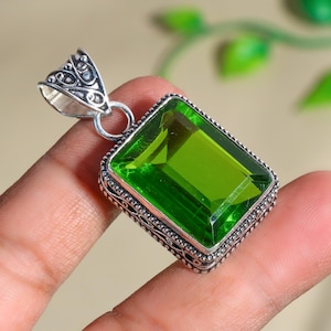 Puede incluir: Un colgante de plata con un diseño ornamentado presenta una gran piedra preciosa rectangular de color verde esmeralda.