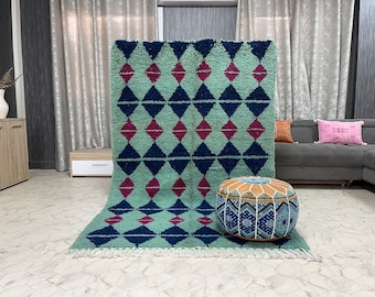 Op de afbeelding: Een groen, blauw en roze geometrisch patroon tapijt met een franje rand. Een ronde, bruin en blauwe gepatroneerde poef staat op het tapijt.