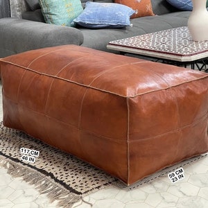 May include: A brown leather ottoman with a square shape and stitched panels. The ottoman measures 46 inches wide, 23 inches deep, and 18 inches high.