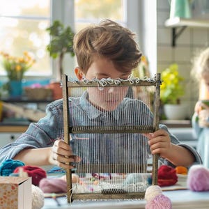 May include: A child is working with a wooden weaving loom, surrounded by colourful yarn and craft materials on a table. Another person is in the background. The setting is a bright kitchen, indicating a crafting activity.