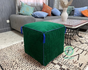 May include: A square, emerald green ottoman with blue trim and tassels. The textured fabric cube sits on a patterned rug, with a gray sofa and decorative pillows in the background. A coffee table and vase are also visible.