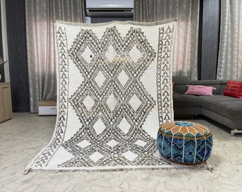 May include: A white and gray Moroccan rug with a geometric pattern. The rug is laid out on a light gray floor with a blue and brown pouf in the foreground.