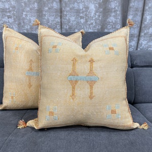 May include: Two beige throw pillows with tassels and embroidered geometric patterns in blue and orange.