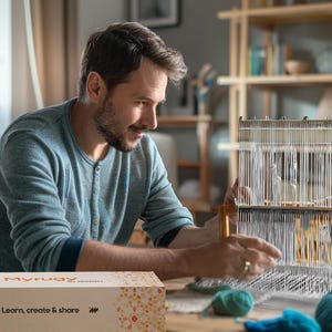 May include: A man and a child are using a wooden weaving loom. The loom has white threads, and colourful yarn balls are on the table. A box with the text "Learn, create & share" is in the foreground, suggesting a craft activity.