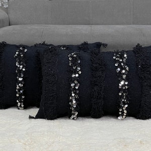 Könnte beinhalten: Drei schwarze Kissen mit silbernen Pailletten und Fransenbesatz. Die Kissen liegen auf einem weißen, flauschigen Teppich.