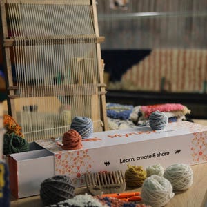 May include: A wooden weaving loom with yarn and a box set on a wooden table. The box has the text "Myrug" and "Learn, create & share". Various coloured yarn balls and a comb are also visible.