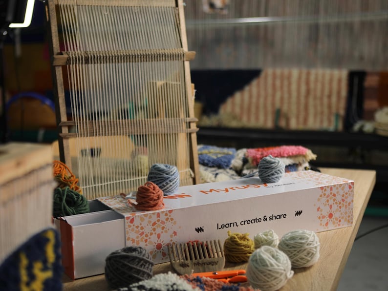 May include: A wooden weaving loom with yarn and a box set on a wooden table. The box has the text "Myrug" and "Learn, create & share". Various coloured yarn balls and a comb are also visible.
