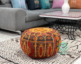 May include: A round, colourful, patterned pouf ottoman with a dark brown background and red, yellow, and orange accents. The pouf is sitting on a white and brown patterned rug.