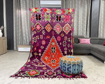 May include: A large, colorful Moroccan rug with intricate geometric patterns in shades of purple, pink, orange, green, and black. The rug is laid out on a tiled floor with a blue and white pouf in the foreground.