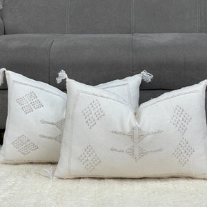 Puede incluir: Dos almohadas blancas con un patrón geométrico en color gris claro. Las almohadas tienen borlas en las esquinas y están dispuestas sobre una alfombra blanca y esponjosa.