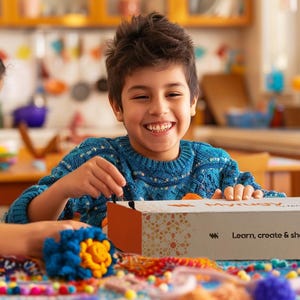 May include: A boy and girl are smiling as they work on a craft project. The boy wears a blue sweater, the girl a grey shirt. A box with "Learn, create & share" sits on the table, alongside yarn and craft supplies, suggesting a creative activity.