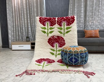 Op de afbeelding: Een wit en crèmekleurig tapijt met een bloemenpatroon met rode, roze en groene bloemen. Het tapijt heeft franjes aan de randen en wordt getoond op een betegelde vloer met een blauw en wit gepatroneerde poef op de voorgrond.