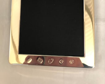 May include: A silver picture frame with a black mat and a polished finish. The frame has a rectangular shape and a wide border. There are five hallmarks on the bottom of the frame.