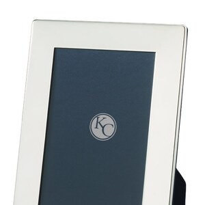 Puede incluir: Un marco de fotos plateado con un borde biselado y un respaldo negro. El marco tiene un pequeño logotipo circular con las letras "KC" en el centro.