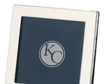 May include: A silver-toned picture frame with a white mat and a blue background. The frame has a raised edge and a small circle with the letters "KC" in the center.