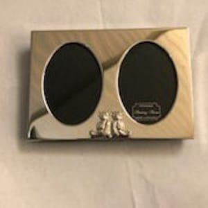 以下が含まれることがあります： 銀製のダブルフォトフレームで、各写真に楕円形の開口部があります。フレームには、手を繋いだ2人の人物の小さなエンボス加工のデザインが施されています。