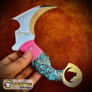 May include: A curved knife with a silver blade and a handle featuring pink and turquoise stone inlays. The handle has a gold-colored end with a bear silhouette. The knife is held in a hand, with the text "Solo Projects Knife Supply" visible.