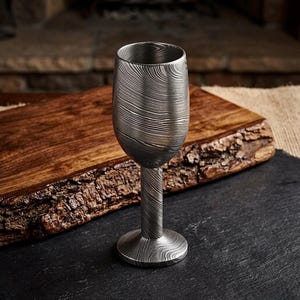 Puede incluir: Una copa de vino de metal artesanal única, con un patrón texturizado. La cristalería tiene un cuenco ancho y un tallo delgado. La copa es de metal plateado y se apoya sobre una superficie oscura. Ideal para vino.