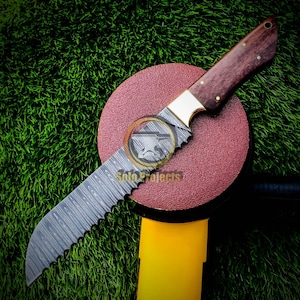 May include: A Damascus steel knife with a serrated edge and a wooden handle. The knife has a brass bolster and is displayed on a red circular surface. The text "Solo Projects" is visible on the blade.
