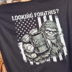 May include: Black t-shirt with a graphic of a smiling Bigfoot holding a 10mm socket wrench in front of an American flag design. The text "LOOKING FOR THIS?" is printed above the flag.