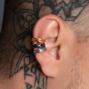 Puede incluir: Tres puños de oreja en oro, negro y plata se usan en la oreja. Los puños de oreja tienen un diseño trenzado. La persona tiene un tatuaje en la nuca y en el brazo.