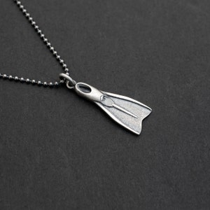 Puede incluir: Un collar de plata con un colgante de aleta de plata. La aleta es un diseño detallado en tres dimensiones.