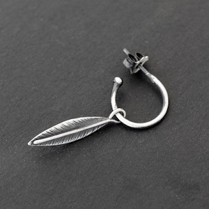 Peut inclure: Une boucle d'oreille en argent avec un petit pendentif en forme de plume. La boucle d'oreille est sur un fond gris foncé.