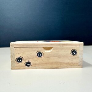 Op de afbeelding: Een lichtgekleurde houten kist met een schuifdeksel. De kist is versierd met zwarte roetgeesten met witte ogen. De kist heeft een kleine, gebogen inkeping om het deksel te openen. De achtergrond is donkerblauw.