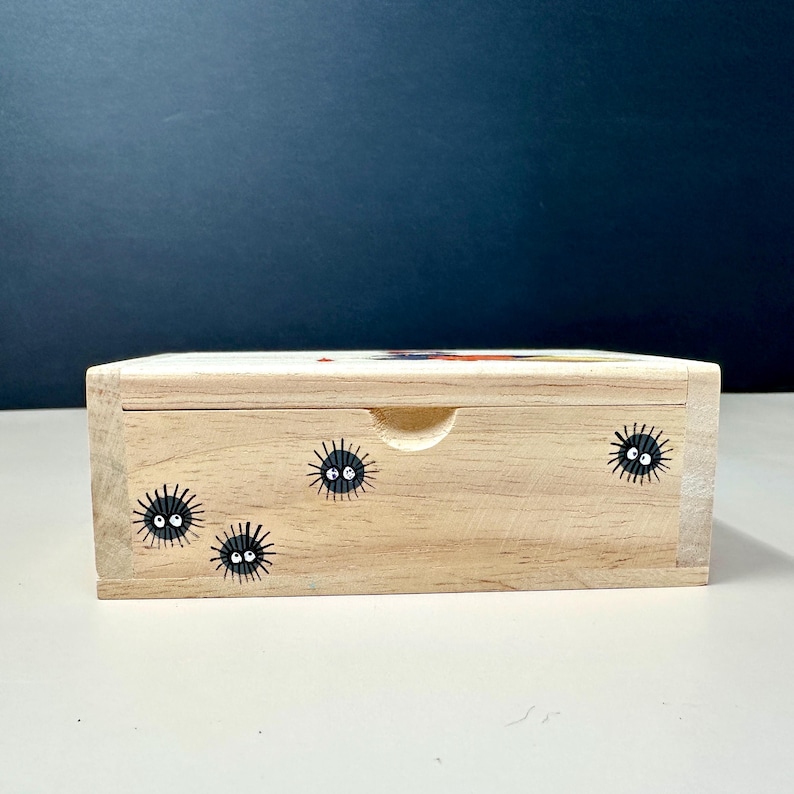Op de afbeelding: Een lichtgekleurde houten kist met een schuifdeksel. De kist is versierd met zwarte roetgeesten met witte ogen. De kist heeft een kleine, gebogen inkeping om het deksel te openen. De achtergrond is donkerblauw.