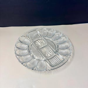 May include: Clear glass serving platter with multiple compartments. The platter has a central section with three rectangular compartments and a surrounding ring of individual sections. The edges are scalloped, and the glass has a textured, patterned design.