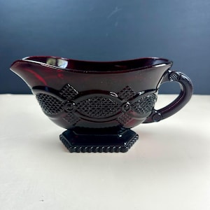 May include: A deep red glass gravy boat featuring a textured diamond pattern across the body, a curved handle, and a scalloped base. The translucent vessel has a wide pouring spout and a repeating geometric design embossed on the exterior surface.