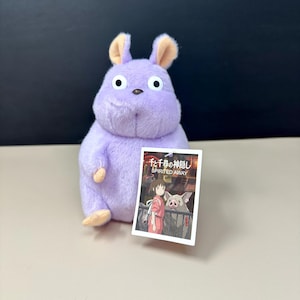 May include: A lavender plush toy resembling a character from "Spirited Away", with large eyes and tan-coloured ears and paws. It holds a tag with artwork from the film. The toy is designed to be a soft, cuddly item.