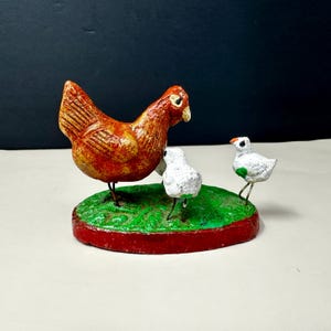 May include: A handcrafted chicken figurine with two chicks on a green base with a red border. The hen is brown and the chicks are white with green accents. The figurine is a charming decorative piece.