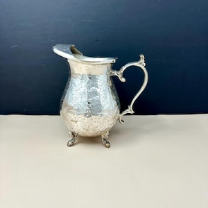 May include: A silver-colored metal creamer pitcher featuring an ornate floral engraving across its rounded body. The vessel includes a curved handle, a flared pouring spout, and stands on three small, decorative feet with a polished finish.