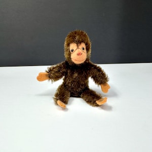 May include: A soft toy monkey with brown fur, tan hands and feet, and a light brown face. The monkey has a pink nose and is sitting on a white surface with a black backdrop.